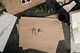 Brown rectangular pieces of paper sit askance in a pile in the center with the top one reading the word, “no” in black inked calligraphy. Surrounding the pieces of paper are various art and office materials sitting atop a gray marbled countertop.