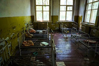 An abandoned room with peeling yellow and white paint on the walls contains several small metal bed frames, many with missing or damaged mattresses. Torn sheets, stuffed dolls, and scattered papers lie on the beds and dusty wooden floor. Light filters through large, grimy windows, casting a dim glow over the scene. The room feels still and long-forgotten, with signs of past use by children.