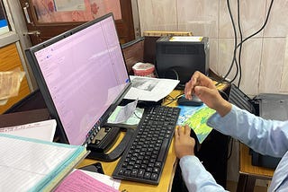 An office setting where one person is extending money towards the other, who is sitting in front of a computer, suggesting a payment or service environment.