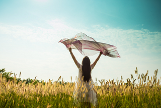 Woman standing in a field in a long dress holding a scarf above her head in the breeze