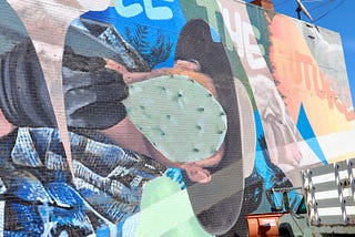 Murial, of a man holding a cactus leaf up to his face dressed like a field worker by the words “See The Future”, by a truck.