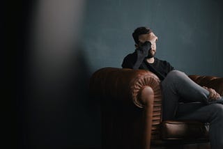 Dark photo of a man sitting on a leather couch with his head in his hands.