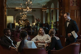 A server at a fancy restaurant dressed in a suit addresses a group of four people sitting around a table