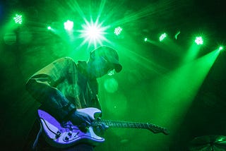 Cox in ballcap playing guitar under the glare of a green stagelight.