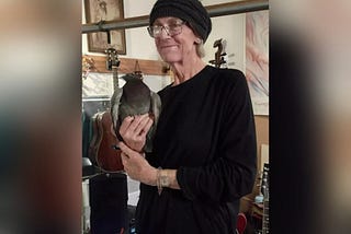 tall older white woman wearing a black top and a black knit hat. she’s holding a pigeon. she has a half smile.