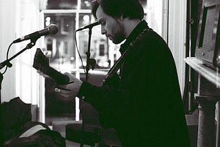 A black and white picture of a young man (Seth Elton) in a small recording studio playing an acoustic guitar. There is a microphone in front of him, waiting expectantly for him to sing into it.