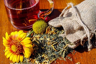 Dried herbs and cut flowers spill out of a hessian bag beside a cup — organic tea