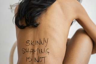 A slender woman, knees to her chest, with writing on her back reading: “skinny shaming is not okay.”