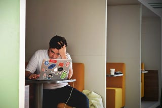 Young man sitting in a booth looking at his laptop with a hand on his head, visibly frustrated.