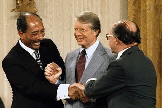 Color photograph of the three men — Egyptian President Anwar Sadat (left), U.S. President Jimmy Carter (center), and Israeli Prime Minister Menachem Begin (right) — in a standing position, shaking hands and smiling.