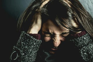 Headshot of a woman in a dark green sweater holding her head while scrunching her forehead, eyes, and nose as if in anger or frustration.