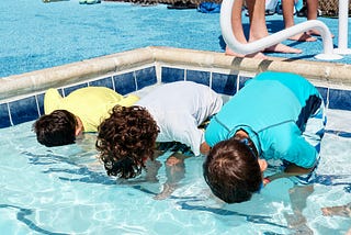 Three young boys stand side-by-side on a step in a public swimming pool with their faces in the water. From left, the youngest is wearing a yellow rash shirt, the middle boy is wearing white, and the eldest boy on the right is wearing a blue rash shirt. They all have brown hair, and the middle boy has curls. Behind them, standing at the edge of the pool, are two more sets of feet.