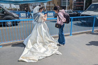 A woman standing with her back to us wearing a wedding dress and her assistant standing next to her near a railing by a busy road.