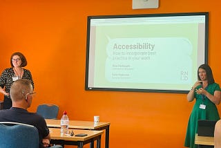 Two women stand on either side of a screen that says ‘Accessibility: how to incorporate best practice in your work. Mhari McNaught, Interaction Designer, Katie Dickerson, Service Designer’. The woman on the right is doing BSL interpretation. There is a man sitting who is sitting in front of a table and looking at the screen who is wearing a hearing aid.