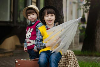 Two dark-haired boys with hats sit outside in the rain. They have a suitcase and an umbrella.