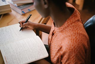 A Black woman writes with a pen in a notebook at a desk