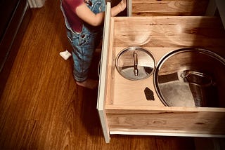 Image shows an open kitchen drawer with two pot lids and one small rock inside of it. Next to the drawer is a small child in overalls.