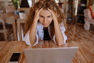 A woman with curly hair holds her head in frustration while looking at a laptop.