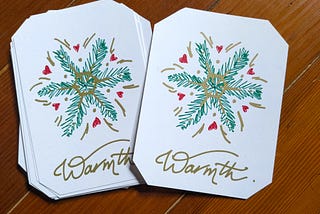 Two white cards laying on brown floorboards, with their corners cut off; the left one sits atop a small stack. The paper shows a “snowflake” design, made of green fir sprigs, red hearts, and gold embellishments. Underneath is “Warmth.” written in gold script.