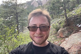 The author is standing in front of mountains, some trees, and rocks. He’s smiling. He has short brown hair and a brown beard. He’s wearing sunglasses.