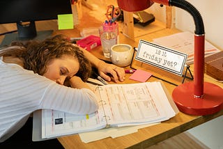 A woman asleep with her head on her desk and her elbow on an open notebook.