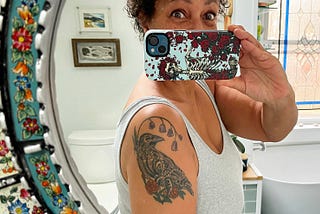 Selfie taken in my bathroom mirror, phone covers my lower face but my eyes look happily surprised. Wearing a grey tanktop that reveals my tattoo of a crow with flowers.