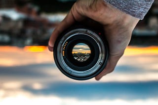 Image of a camera lens up against a blurry landscape, with the inner eye of the lens reflecting a clear landscape.