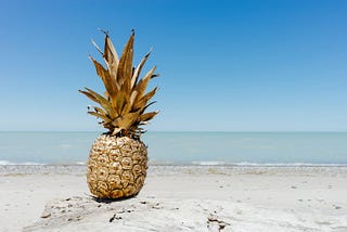 A gold-painted pineapple, sitting on a beach, overlooking blue waters and a blue sky.
