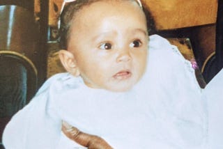 A photograph of me (Kashala, aged around 6 months or so) as a baby, dressed in white, waiting to get christened.