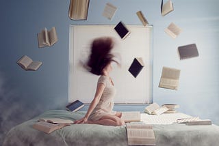 A girl sits on her legs in her bed, books flying around her.