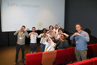 Nine people, making heart shapes with their hands, are grouped in front of a cinema screen that has the words: “Let’s celebrate!”