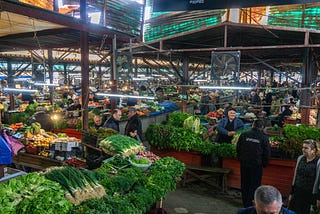 A view of a covered market hall in Kutaisi, Georgia. There are displays of fresh veggies along with vendors and shoppers.