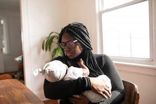 A black woman with braids and glasses holding a baby and gazing at it warmly