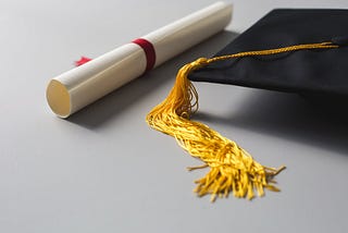 An image displaying a rolled up diploma and a graduation cap on a whitish-grey background.