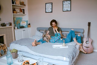 A stock photo of a white, heterosexual couple on a bed. The bed has grey sheets, and the woman has her head in the man’s lap and is feeding him a slice of pizza. At the end of the bed, the pizza sits on a plate with the single slice missing.