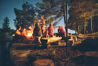 group of people sit around a campfire talking in a forest at dusk