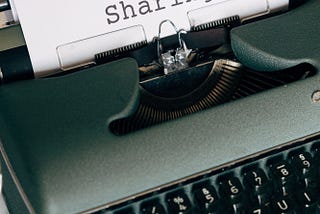 An antique typewriter with a white piece of paper in it that says the word SHARING
