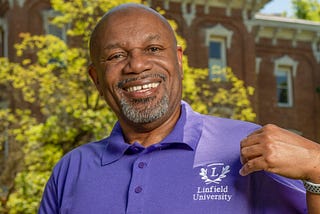 A man points to a logo on his left breast on a purple polo shirt. He is standing in front of a brick building.