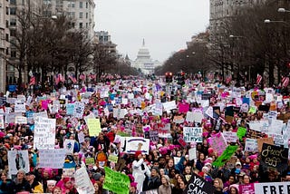 A large crowd of people gathered in a peaceful demonstration in an urban area with the U.S. Capitol building visible in the background. Many participants are holding colorful signs advocating for various causes, including women’s rights, equality, and justice. The atmosphere is vibrant, with a focus on activism and solidarity. American flags and bare trees line the street, emphasizing the setting as a winter march in Washington, D.C.