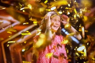 An excited girl in a pink dress in front of a microphone, smiling under a shower of gold confetti
