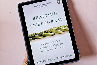 A hand holding an iPad Air tablet tilted slightly to the left, with an image of the book cover of Braiding Sweetgrass by Robin Wall Kimmerer