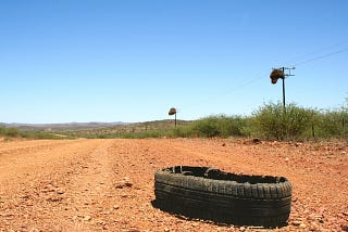 A blown out tire sits on the orange earth of a deserted highway against a backdrop of blue sky and rough, scrubby bushes.