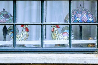 Image: two stained glass rooster sculptures facing each other, with lamps behind each one, viewed through a window pane.