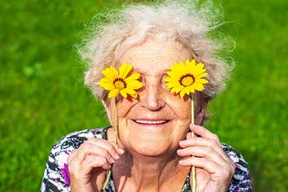 A cheerful grandmother playfully holds yellow daisies up to her eyes.