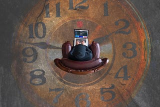 A person sitting on a brown plush sofa with a laptop and with a clock painting on the floor right below where he is sitting