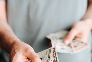 Person in gray t-shirt offering you two cash bills.