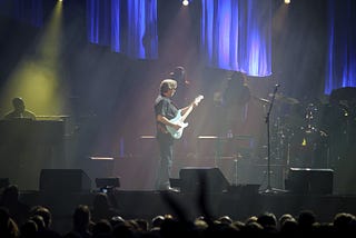 Guitarist Eric Clapton standing center stage playing a white electric guitar.