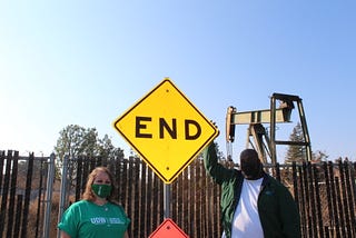 Two union members stand in front of an oil pump and a bright-yellow sign that says “END.”