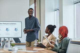 Data Analyst standing next to a screen with pie charts, presenting in a conference room with two people seated at the table.