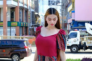 A woman wearing red Filipina Dress standing against a crowded San Pedro Street with shop houses and vehicles in the background.